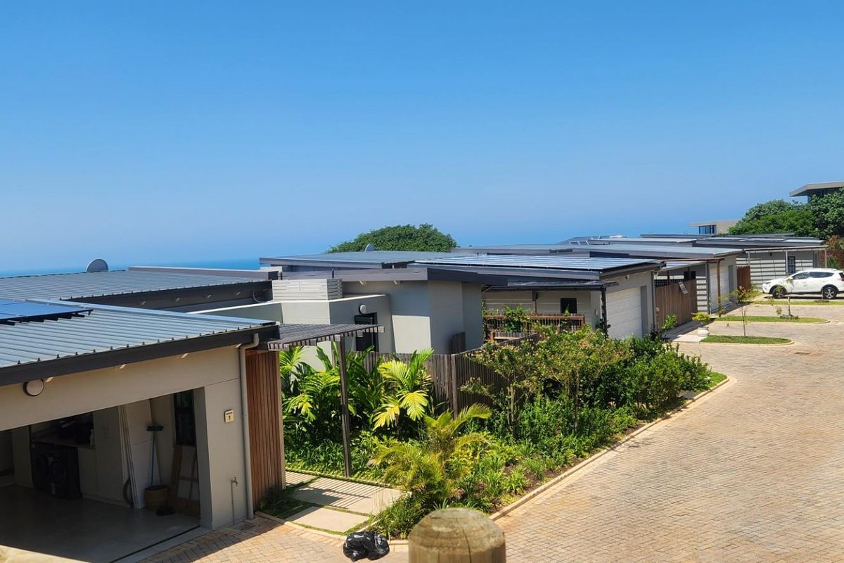 An elevated view of a modern residential complex featuring paved driveways, garages, and solar panels, with a scenic coastal backdrop.