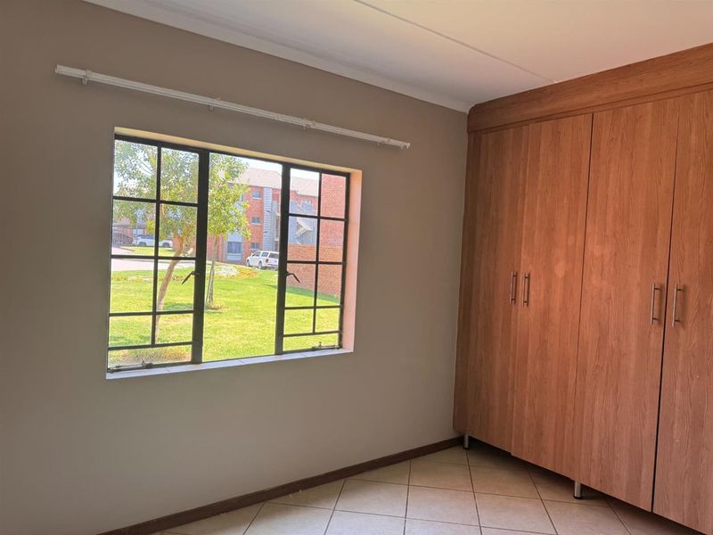 A bright, empty bedroom featuring tiled flooring, built-in wooden cupboards, and a metal-framed window overlooking the complex grounds.