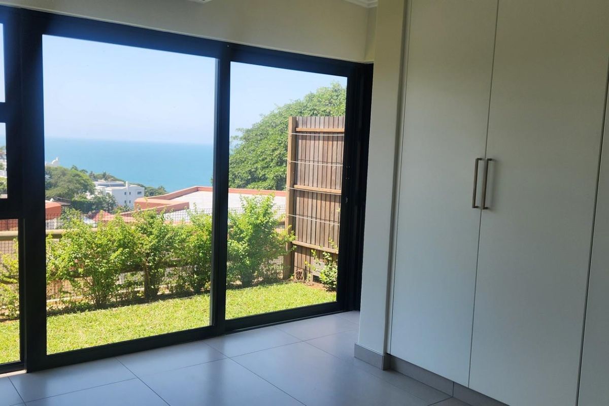 A bright, empty bedroom featuring modern tiled flooring, built-in cupboards, and large sliding glass doors that open to a garden with ocean views.