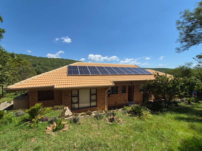 A well-maintained brick house featuring a tiled roof equipped with a large solar panel array, set within a landscaped garden with scenic hillside views.