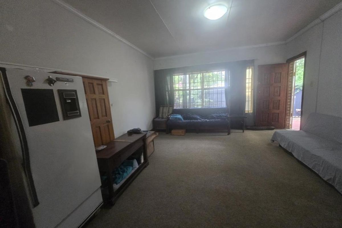 A spacious living area featuring carpet flooring, security bars on the window, and direct access to the exterior through a timber door.