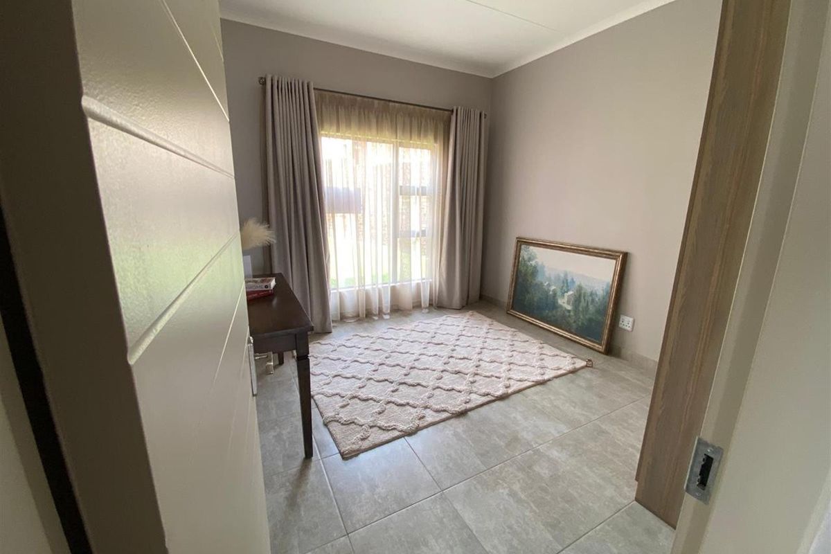 A bright bedroom featuring neutral wall tones, large format floor tiles, and a window dressed with floor-to-ceiling curtains.