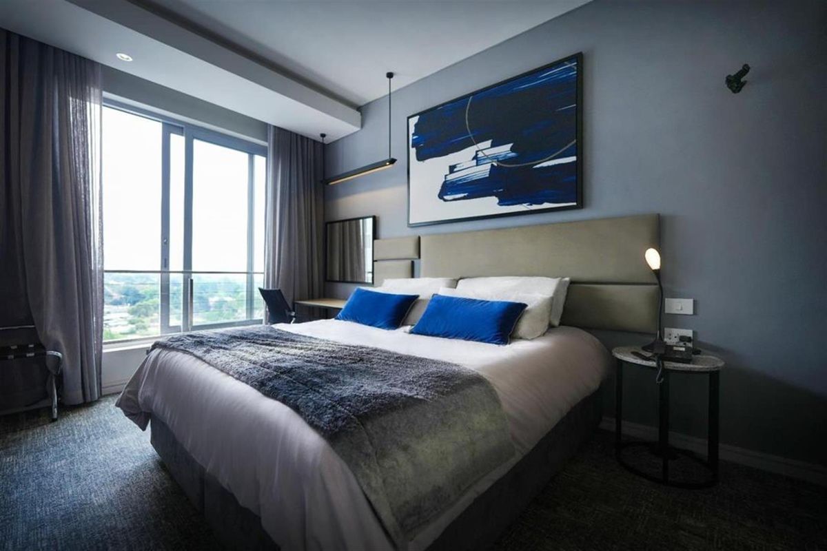 A modern, well-appointed bedroom featuring a built-in upholstered headboard, recessed ceiling lighting, and large sliding glass doors leading to a private balcony.