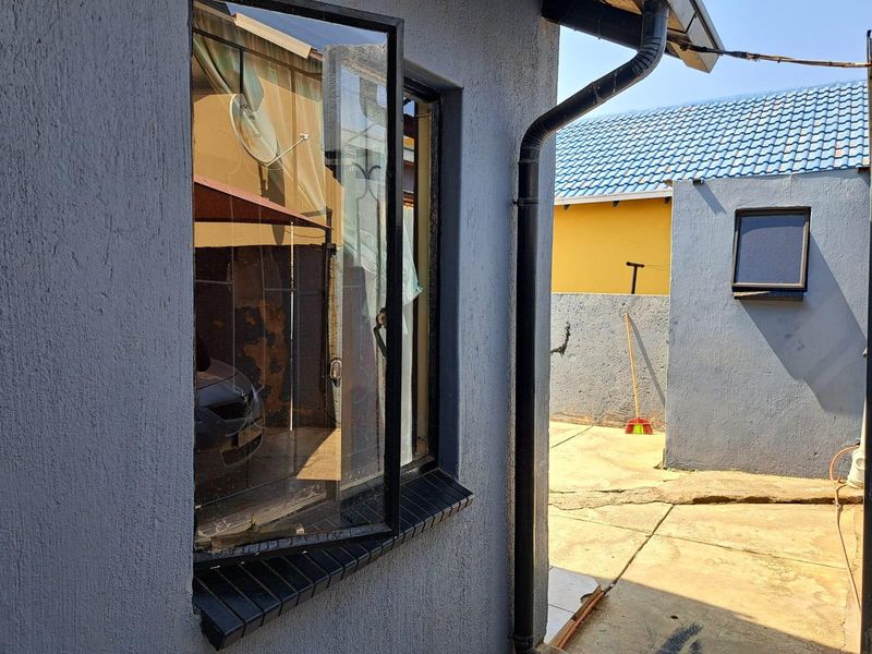 Exterior view of the property showing a side passage with concrete paving, a metal-framed window, and a black downpipe against a textured grey wall.