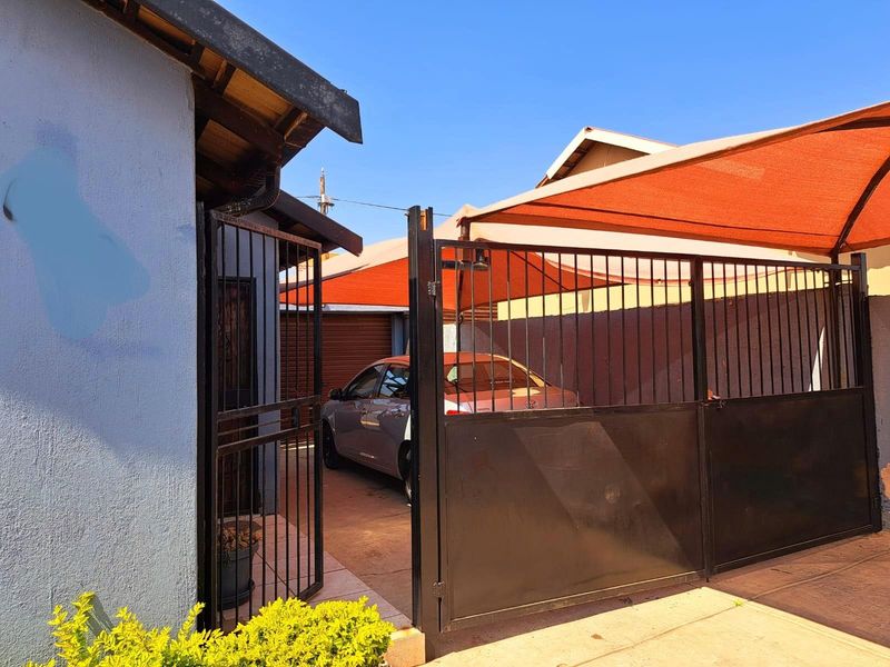 Exterior view of the property entrance featuring a secure metal gate, paved driveway, and a shade cloth carport structure.