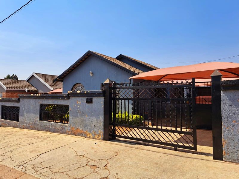Front view of the property featuring a secure metal gate, boundary wall, and a shaded carport area.