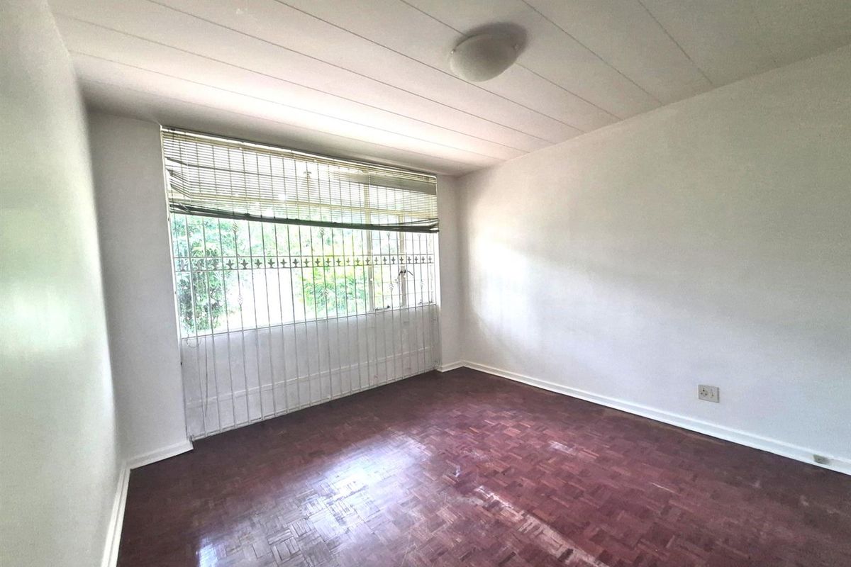 A vacant bedroom featuring traditional parquet flooring, a large window with security bars, and a simple ceiling-mounted light fixture.
