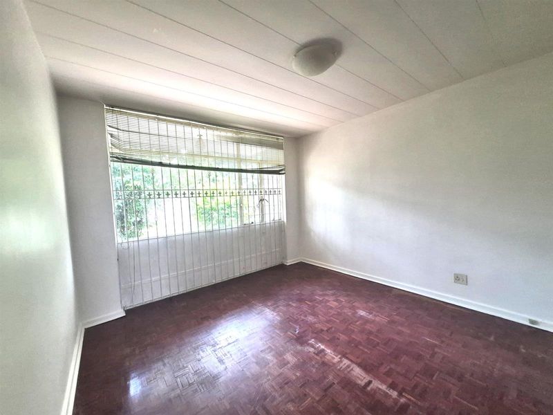 A vacant bedroom featuring traditional parquet flooring, a large window with security bars, and a simple ceiling-mounted light fixture.