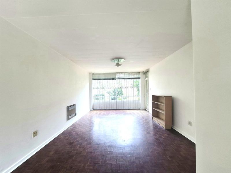 A spacious, sunlit living area featuring classic parquet flooring, a built-in wall heater, and large windows with security bars.