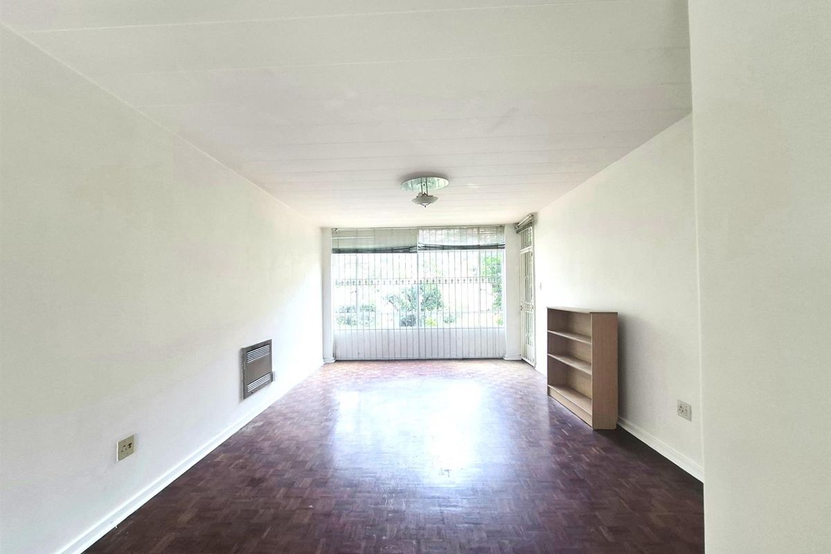 A spacious, sunlit living area featuring classic parquet flooring, a built-in wall heater, and large windows with security bars.