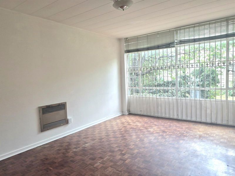 A bright, empty living room featuring original parquet flooring, a built-in wall heater, and large windows secured with decorative metal burglar bars.