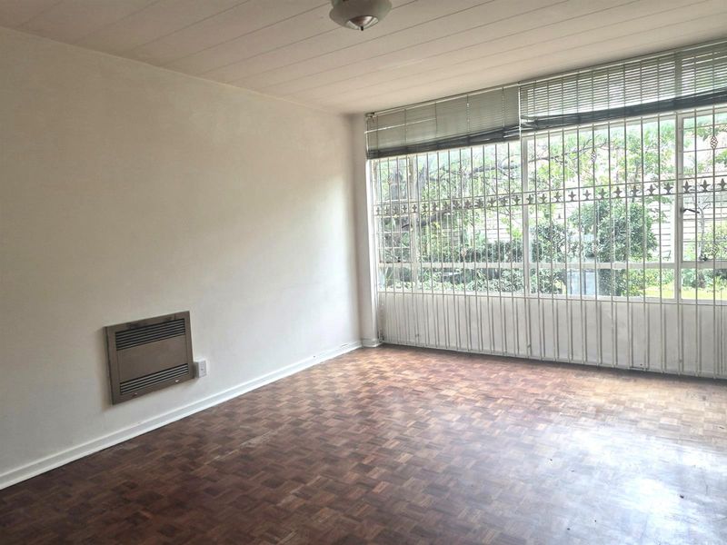 A bright, empty living room featuring original parquet flooring, a built-in wall heater, and large windows secured with decorative metal security bars.
