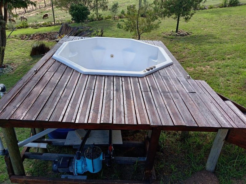 Outdoor octagonal spa bath set into a raised timber deck, featuring exposed pump infrastructure and views over the surrounding farm landscape.