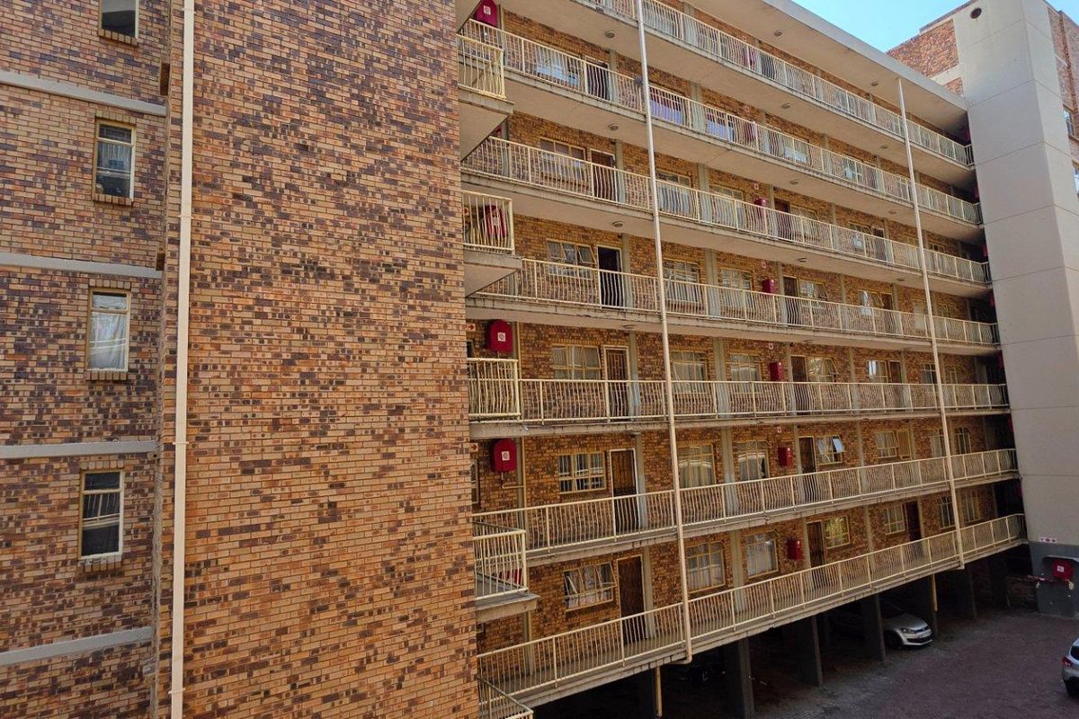 Exterior view of a multi-story apartment building featuring face brick facade, external balcony walkways with metal railings, and ground-level covered parking.