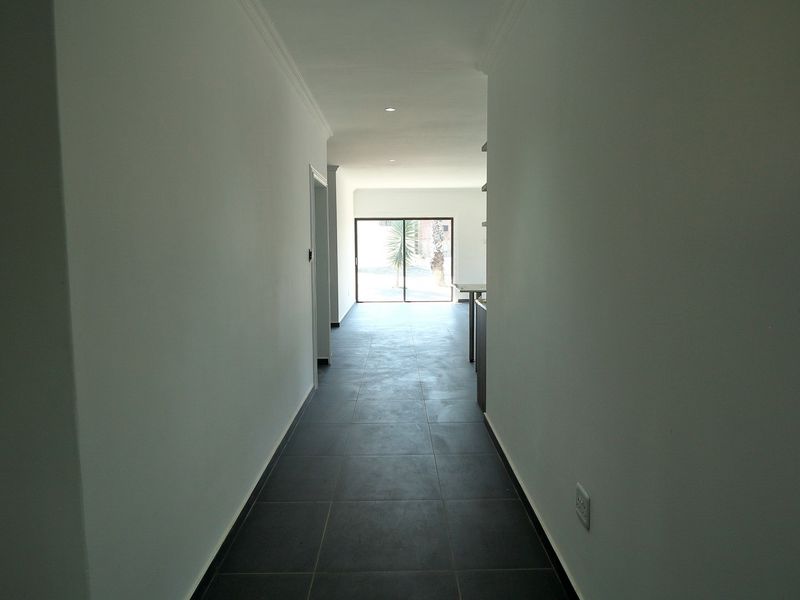 A clean, empty hallway featuring dark tiled flooring and recessed lighting, leading towards a bright living area with a sliding glass door.