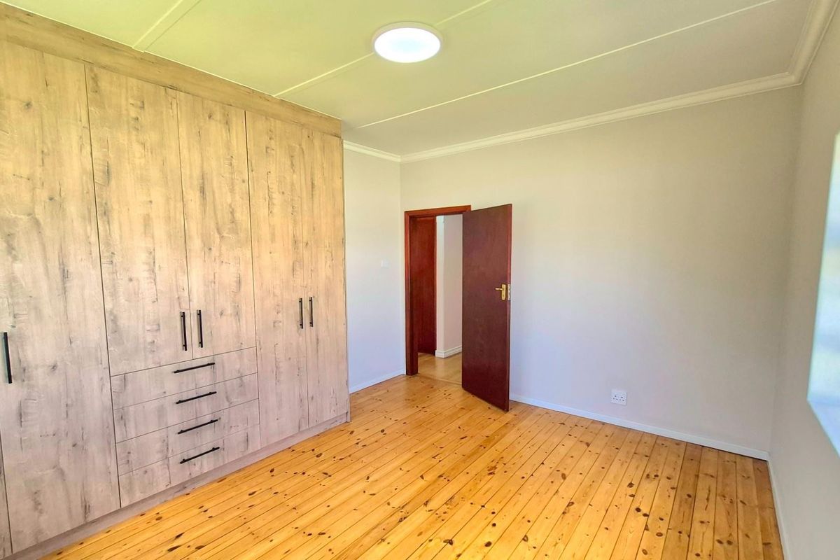 A bright, empty bedroom featuring polished timber flooring, extensive built-in wooden cabinetry, and louvered windows providing natural light.