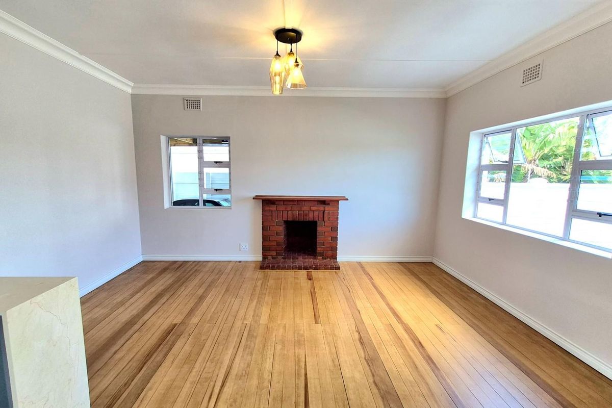A bright, empty living room featuring polished timber flooring, a classic brick fireplace, and large windows providing natural light.