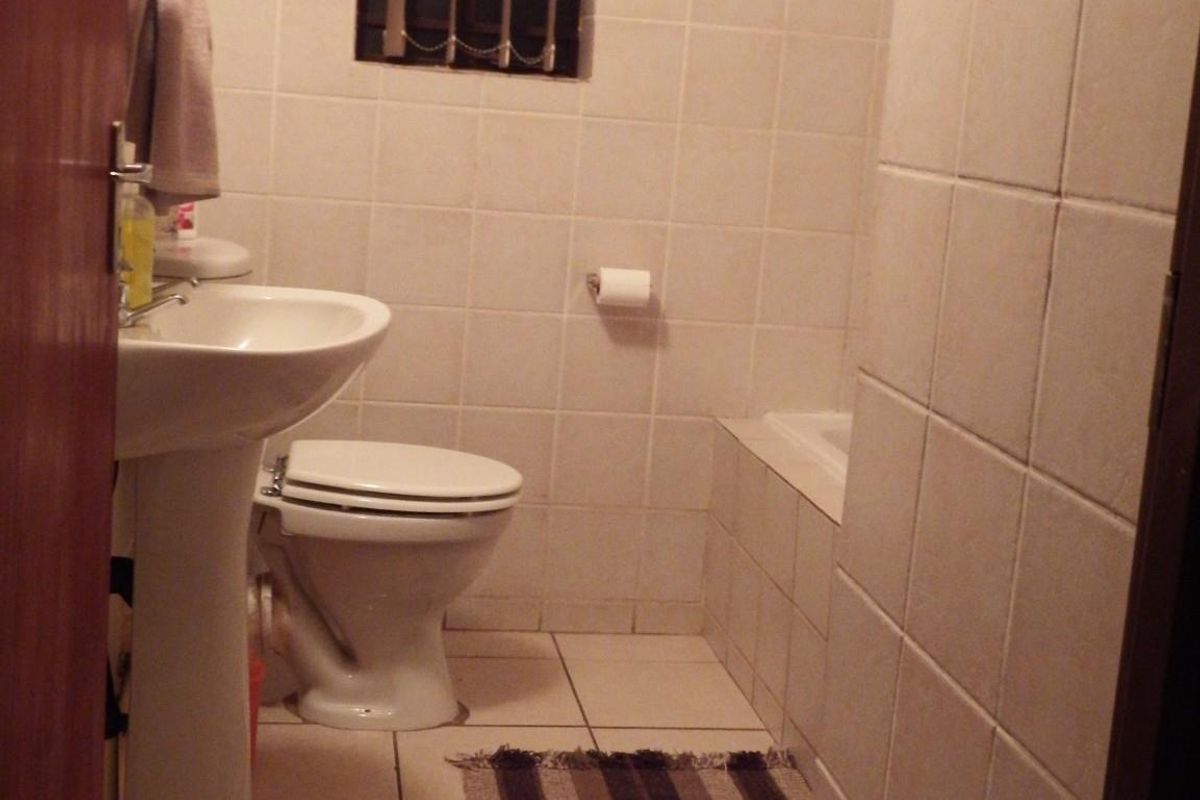 A compact bathroom featuring a pedestal sink, toilet, and tiled bathtub surround with a small window protected by security bars.