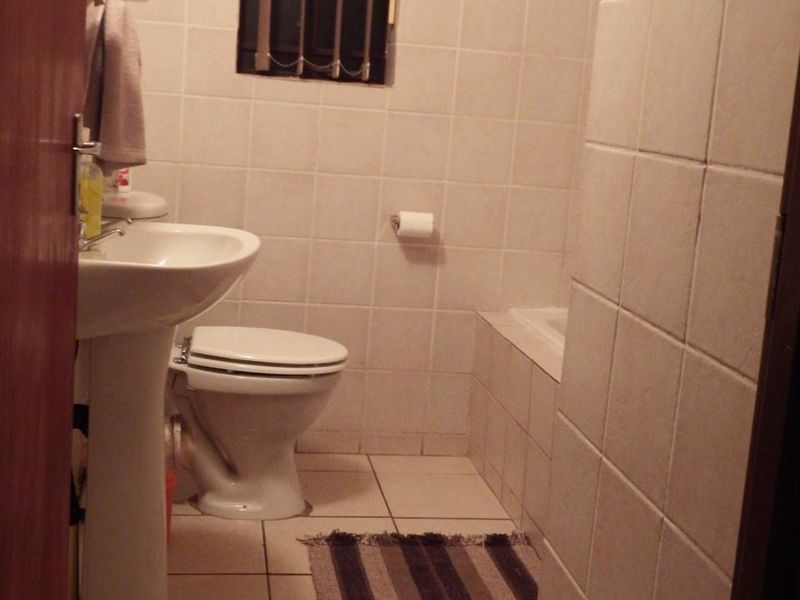 A compact bathroom featuring a pedestal sink, toilet, and tiled bathtub surround with a small window protected by security bars.