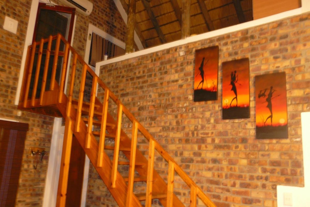 Interior view of a living area featuring exposed brick walls, a wooden staircase leading to a mezzanine level, and a high thatched roof.
