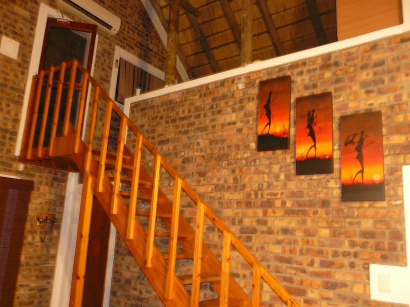 Interior view of a living area featuring exposed brick walls, a wooden staircase leading to a mezzanine level, and a high thatched roof.