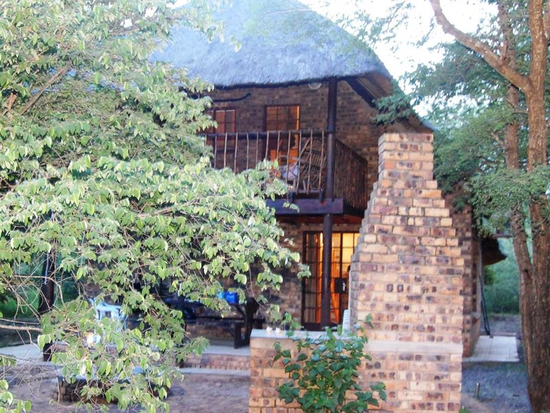 A rustic two-story house featuring a thatched roof, exposed brick exterior, a private balcony, and an outdoor brick fireplace in a natural setting.