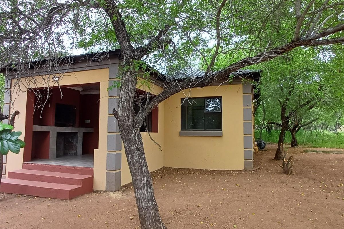 A detached outdoor structure featuring a covered patio with a built-in braai area, set within a natural, tree-filled garden environment.
