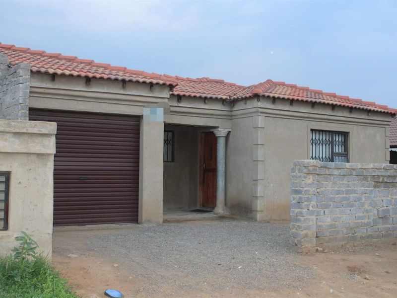 Front exterior view of a single-story house featuring a roller garage door, tiled roof, and an unfinished boundary wall.