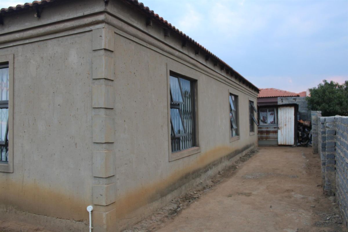 Side view of the house showing the exterior wall, window frames with security bars, and a dirt pathway leading to the rear yard.