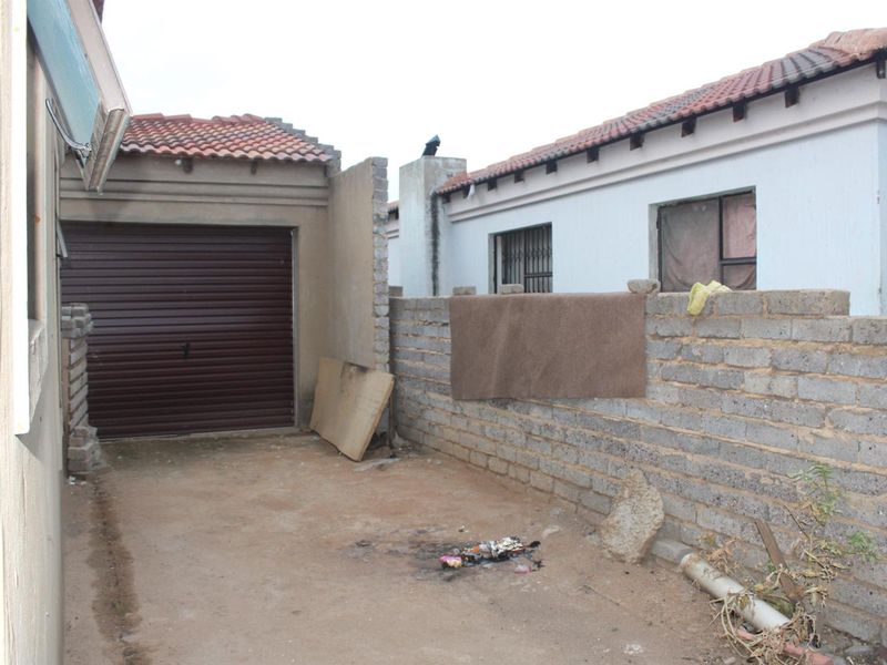 Rear exterior view showing a garage, an unfinished brick boundary wall, and an open paved area requiring maintenance and landscaping.