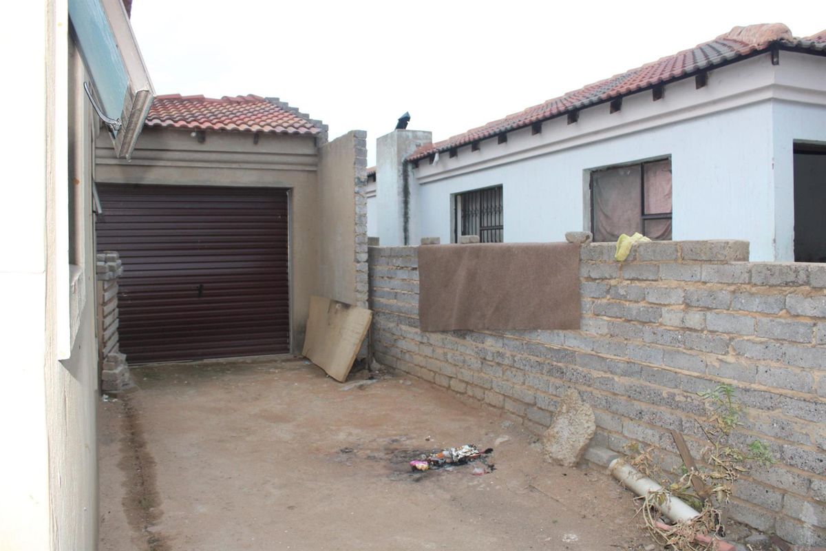Rear exterior view showing a garage, an unfinished brick boundary wall, and an open paved area requiring maintenance and landscaping.