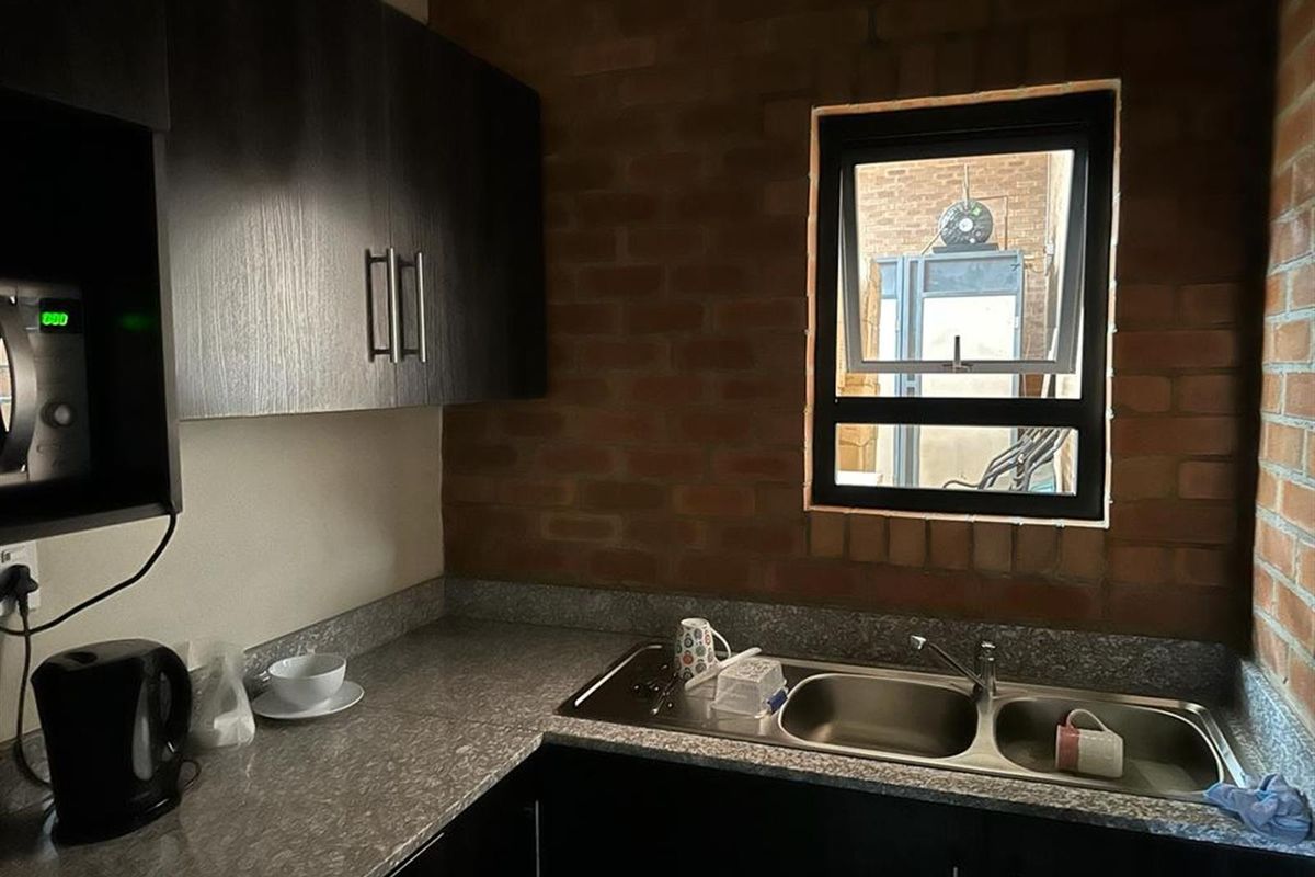 Compact kitchen area featuring dark cabinetry, stone-look countertops, a double stainless steel sink, and an exposed brick feature wall.