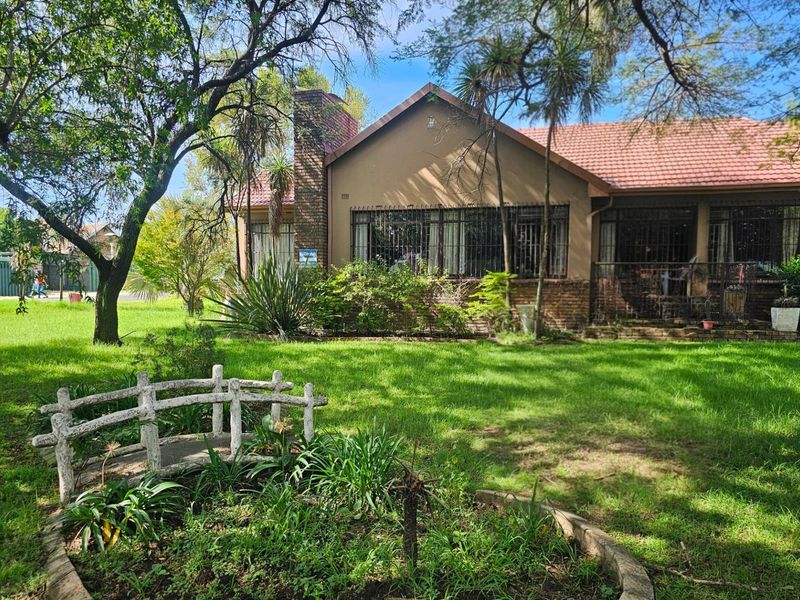 A view of the rear garden featuring a small decorative bridge, mature trees, and the rear elevation of the house with a brick chimney.