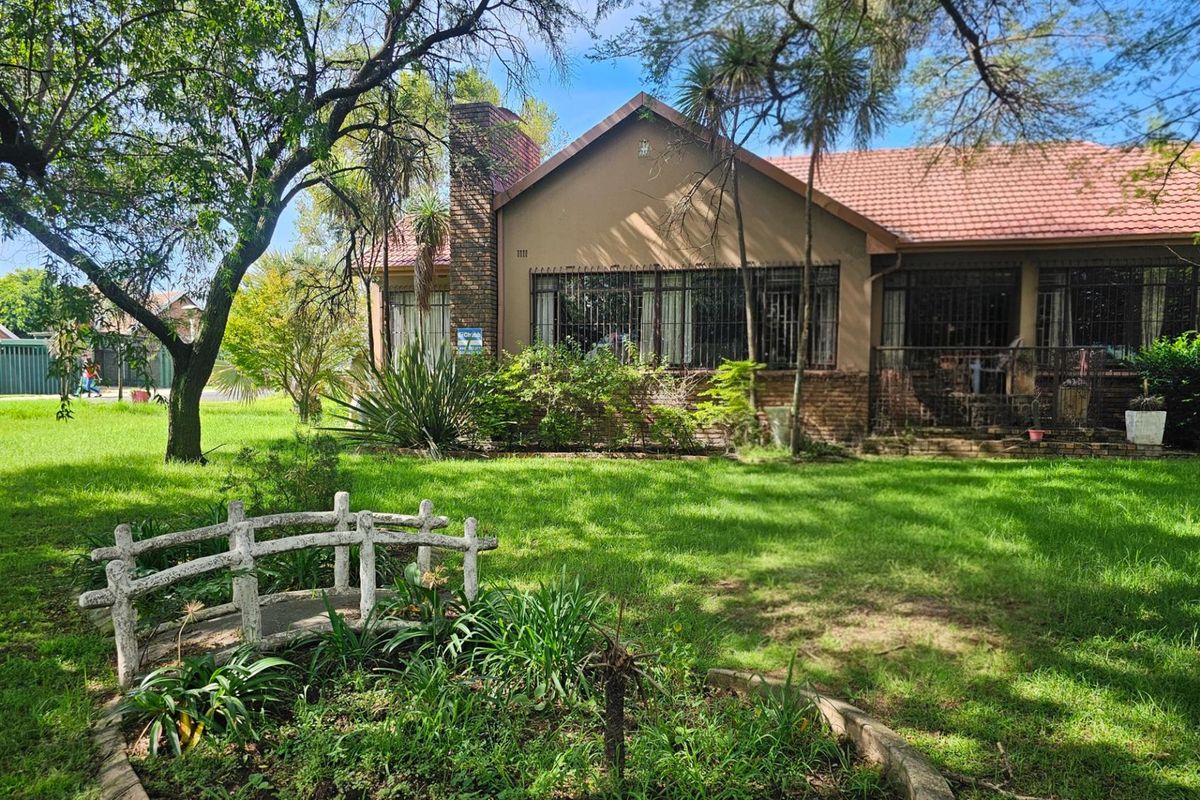 A view of the rear garden featuring a small decorative bridge, mature trees, and the rear elevation of the house with a brick chimney.
