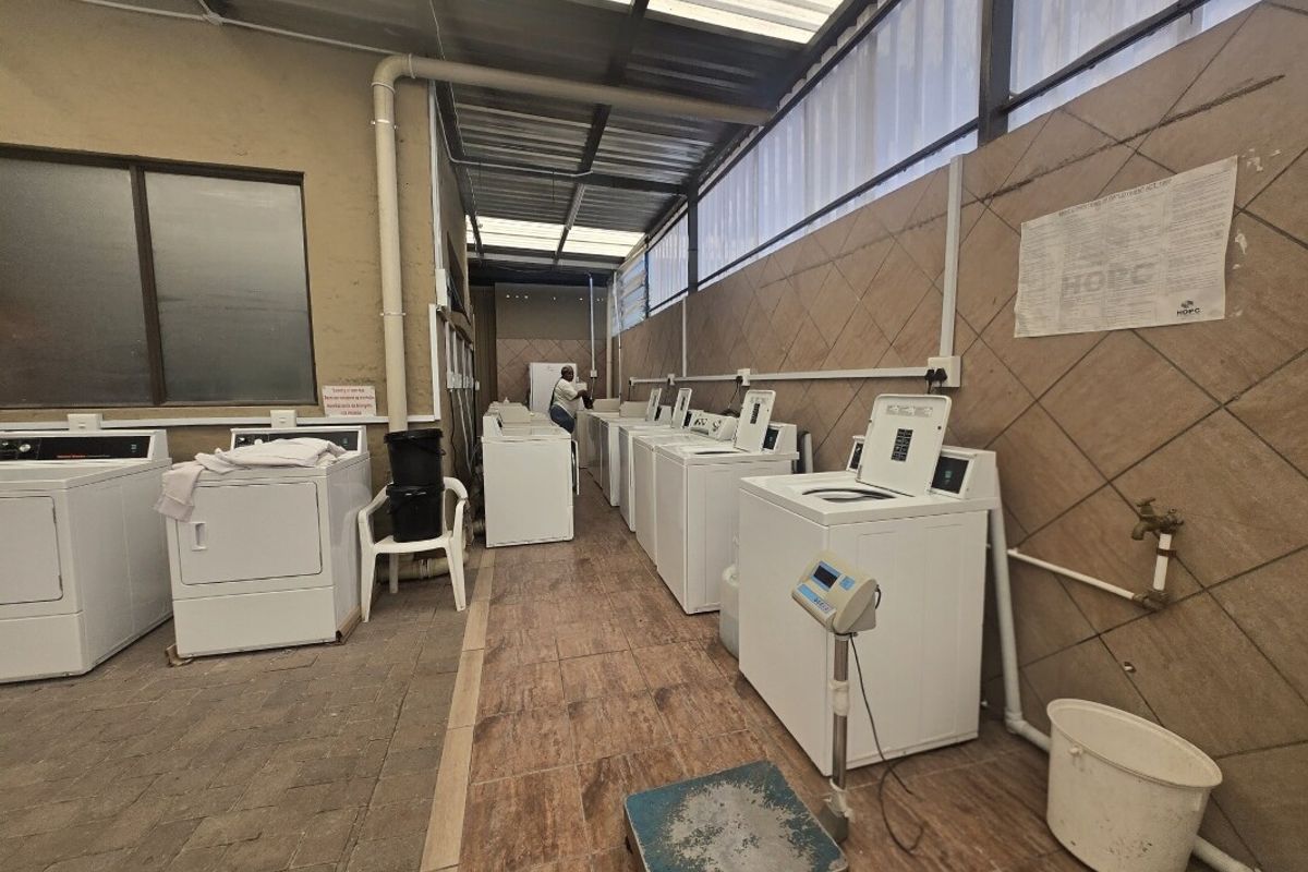 A functional commercial laundry facility featuring multiple washing machines, tiled flooring, and wall-mounted utility connections under a covered structure.
