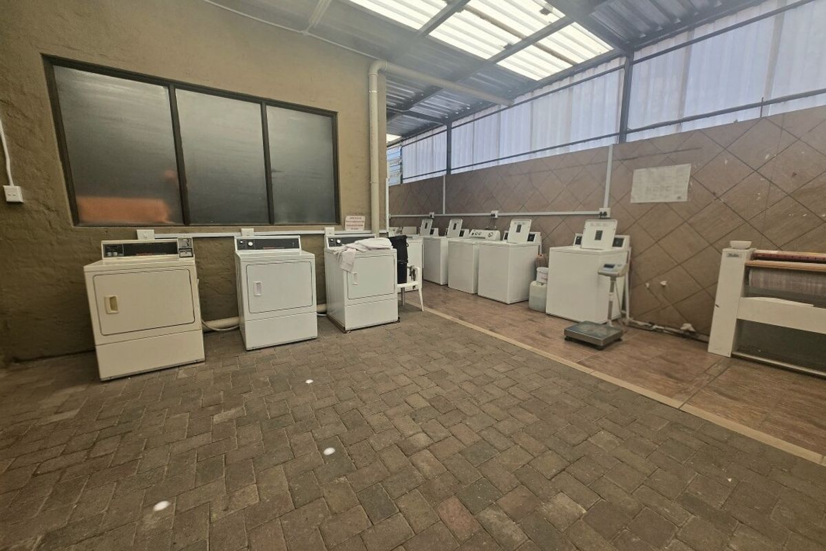A functional commercial laundry area featuring multiple washing machines and dryers, paved flooring, and a covered structure with translucent roofing.