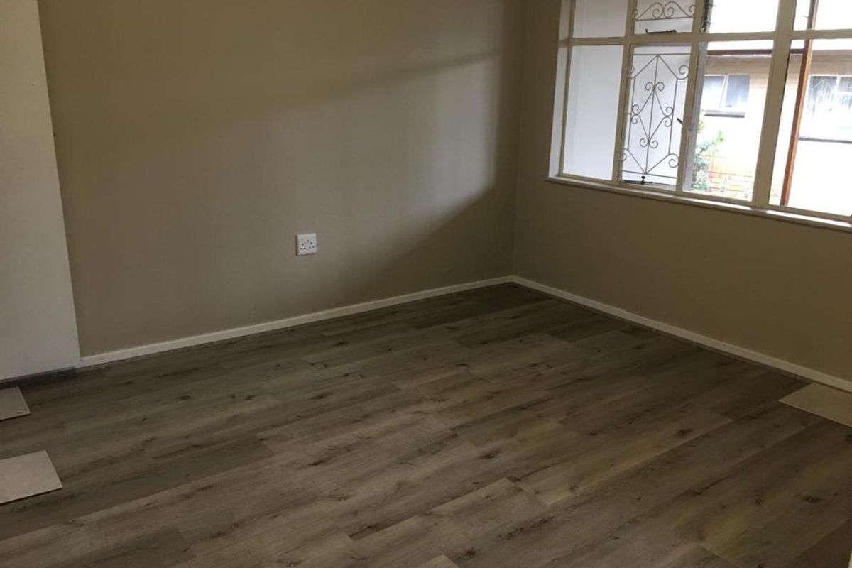 A bright, empty living room featuring modern laminate wood-look flooring and large windows with integrated security bars.
