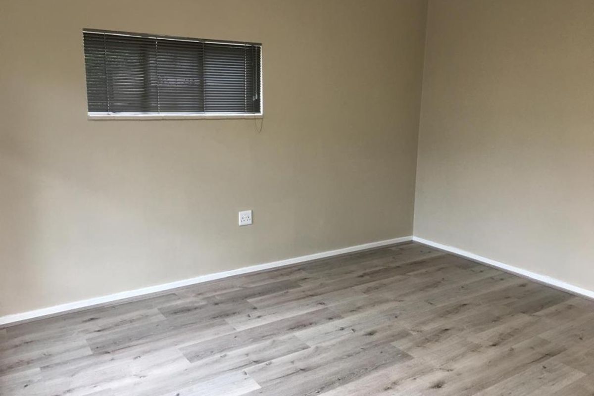 A clean, empty bedroom featuring modern grey-toned laminate flooring, neutral wall paint, and a small window fitted with venetian blinds.
