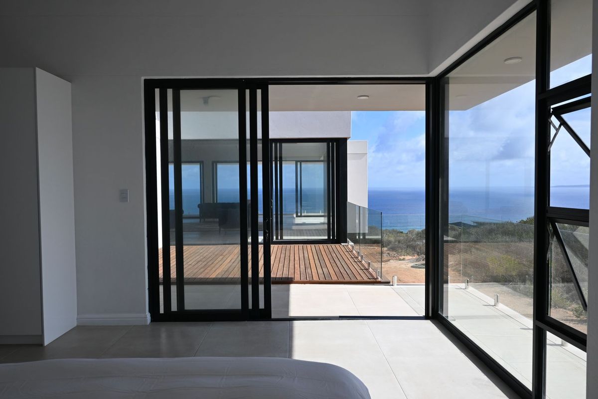 Modern bedroom featuring large format floor tiles, expansive black aluminium sliding doors, and a corner window offering panoramic ocean views and access to a deck.