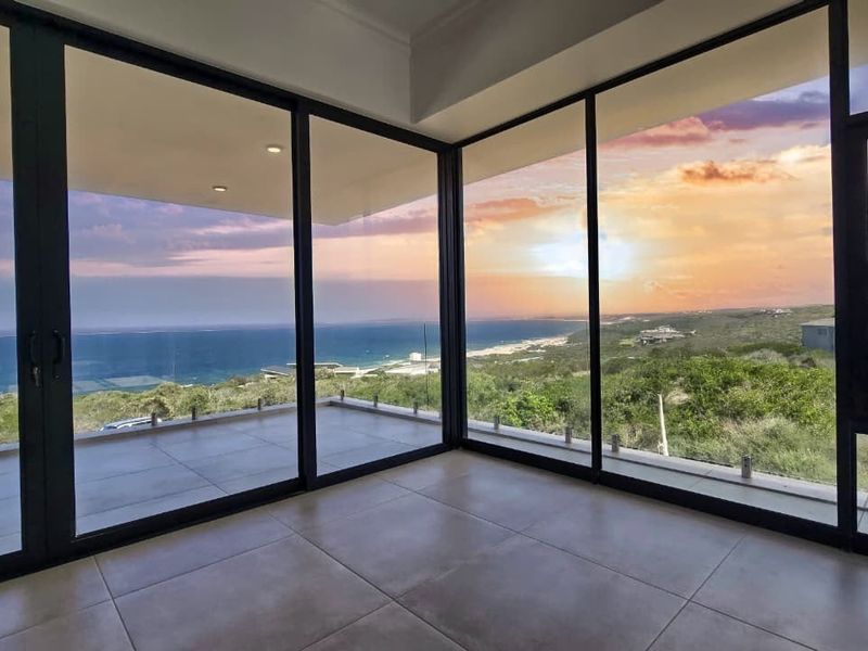 Spacious living area featuring expansive floor-to-ceiling windows and sliding doors that open to a balcony with panoramic ocean and coastal views.