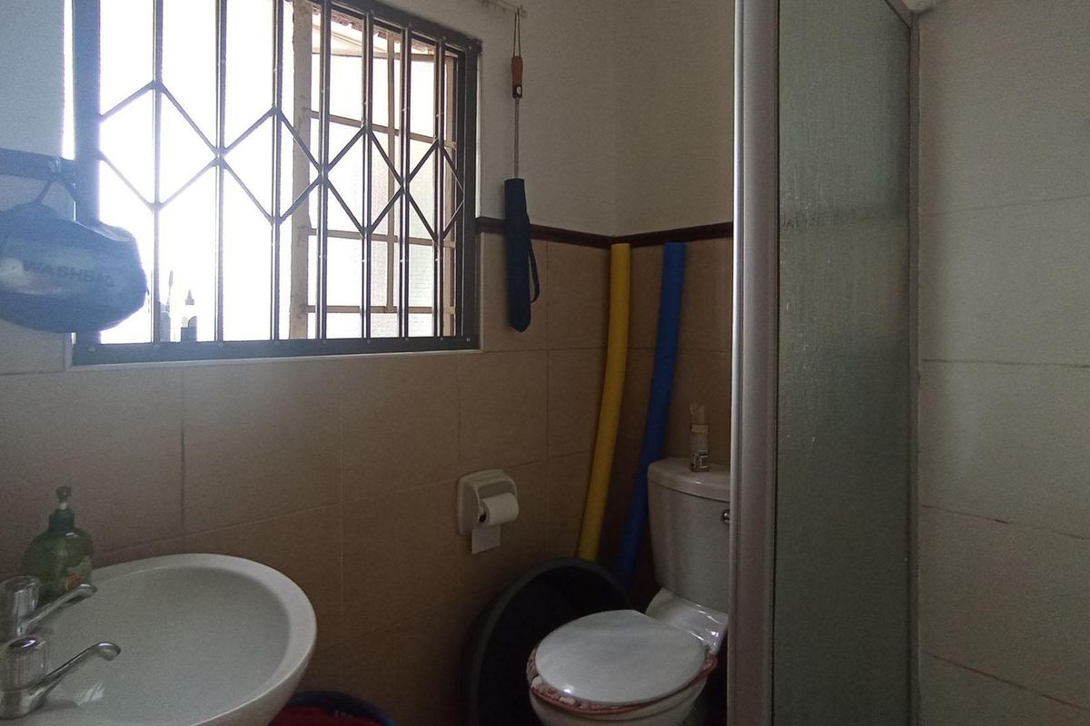 A compact bathroom featuring a shower enclosure, pedestal basin, and toilet, with tiled walls and a window fitted with security bars.