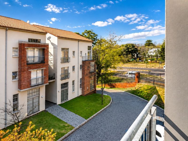 View from a balcony overlooking the apartment complex grounds, featuring a paved driveway, landscaped lawn, and the building's exterior facade.
