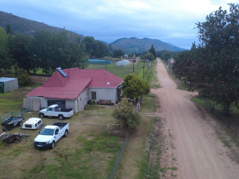 Aerial view of a rural property featuring a house with a metal roof, solar water heater, and surrounding land adjacent to a dirt road.