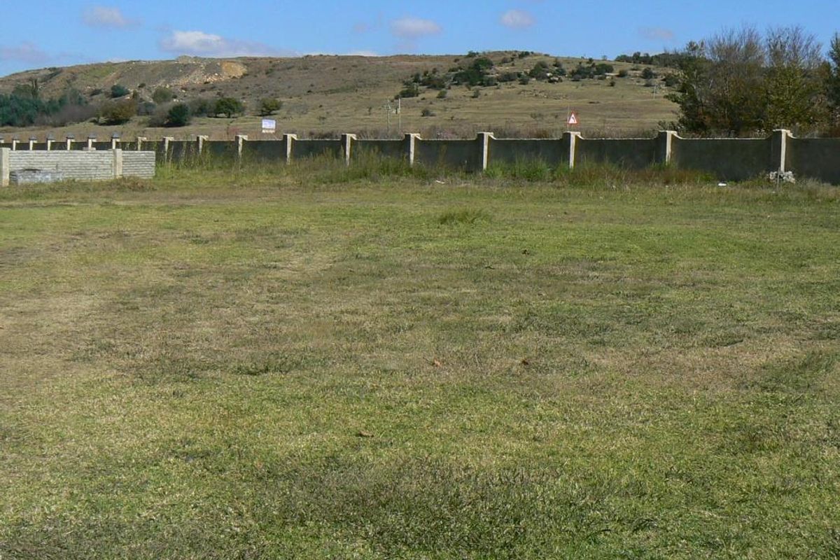 A vacant residential plot featuring a grassed area enclosed by a perimeter wall with concrete fence posts, set against a hilly landscape.