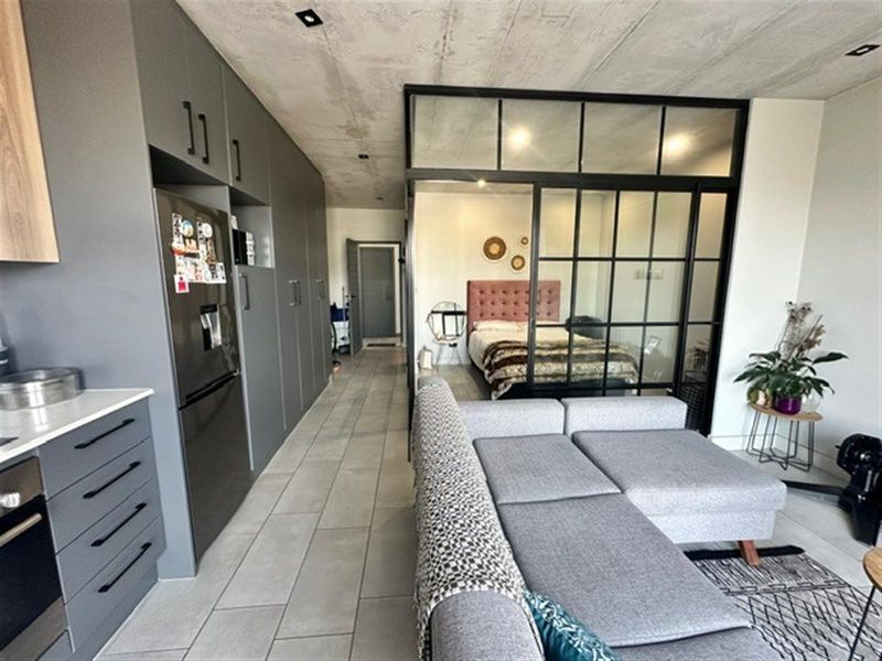 Modern open-plan apartment featuring a sleek grey kitchen, tiled flooring, exposed concrete ceiling, and a stylish industrial-style glass partition separating the bedroom area.