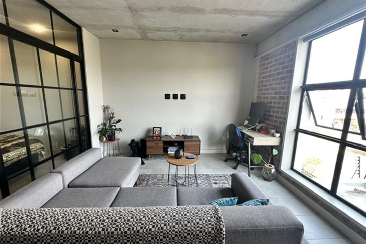 Modern open-plan living area featuring an industrial aesthetic with an exposed concrete ceiling, brick feature wall, and large black-framed glass partition.