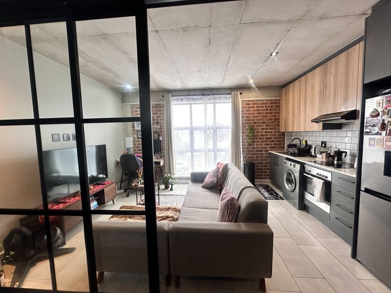 Modern open-plan apartment featuring an exposed concrete ceiling, brick accent wall, and a sleek kitchen with integrated appliances and tiled flooring.