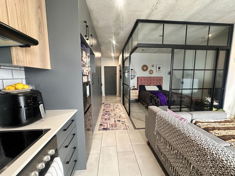 Modern open-plan apartment featuring a sleek kitchen with built-in cabinetry, tiled floors, exposed concrete ceilings, and a stylish glass-partitioned bedroom area.