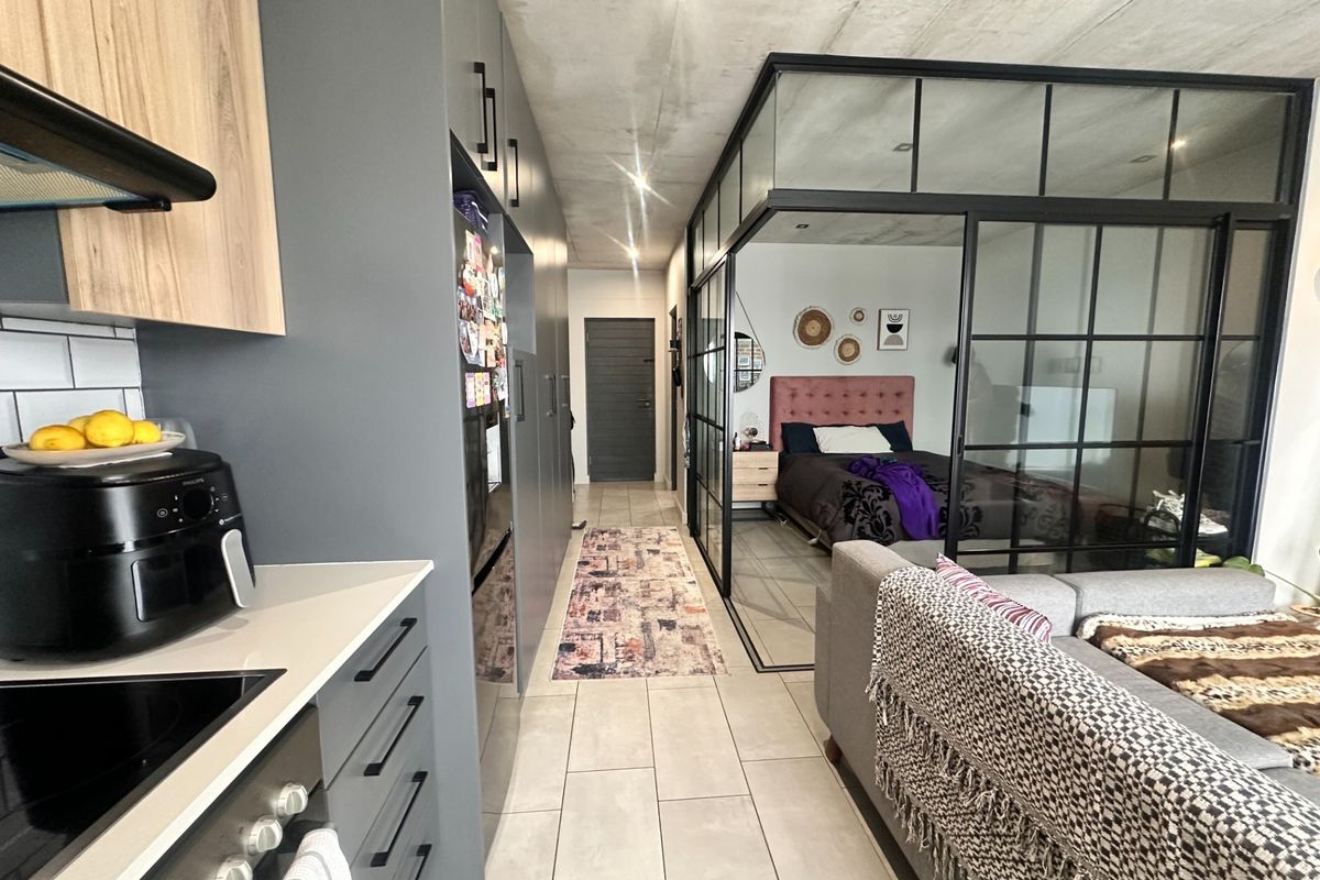 Modern open-plan apartment featuring a sleek kitchen with built-in cabinetry, tiled floors, exposed concrete ceilings, and a stylish glass-partitioned bedroom area.
