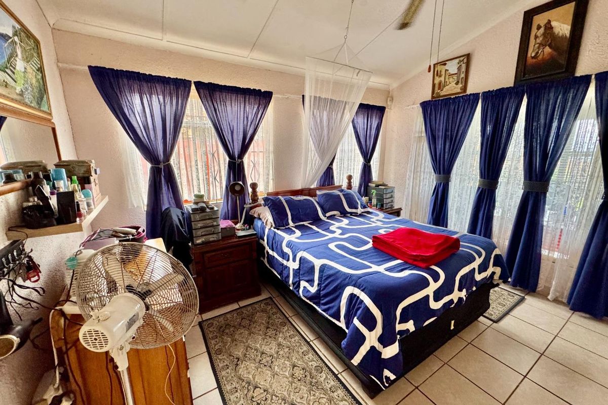A bedroom featuring tiled flooring, a ceiling fan, and large windows with curtains, providing a functional and lived-in space.